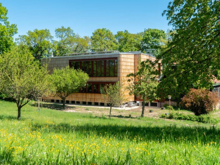 © Alexander Stadler Seitenansicht des neu gebauten Gärtnerhaus der Hochschule für Agrar- und Umweltpädagogik im Sommer umgeben von Bäumen, Sträuchern und Wiese.